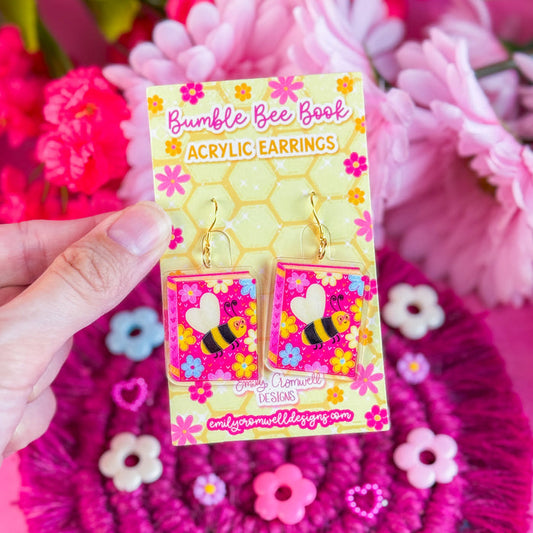 Hand holding a package of 'Bumble Bee Book' acrylic earrings with pink flowers in the background.