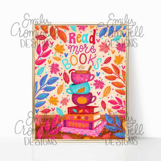A fall art print featuring "read more books" a stack of three coffee mugs on top of 4 books, blue orange and pink leaves and foliage surrounding them. The art print is in a shiny gold frame against a white background.