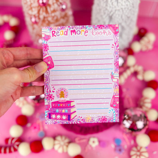 Hand holding a notepad with the words "read more books" at the top and a stack of three pink and purple books in the bottom left corner.