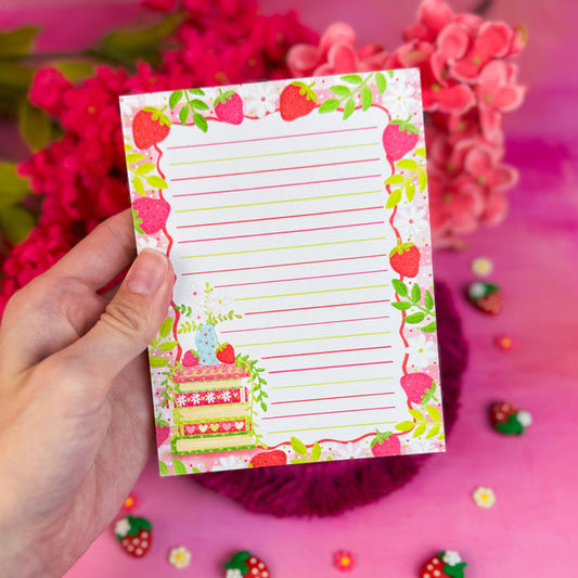 Hand holding a colorful notepad with strawberry design against a pink floral background