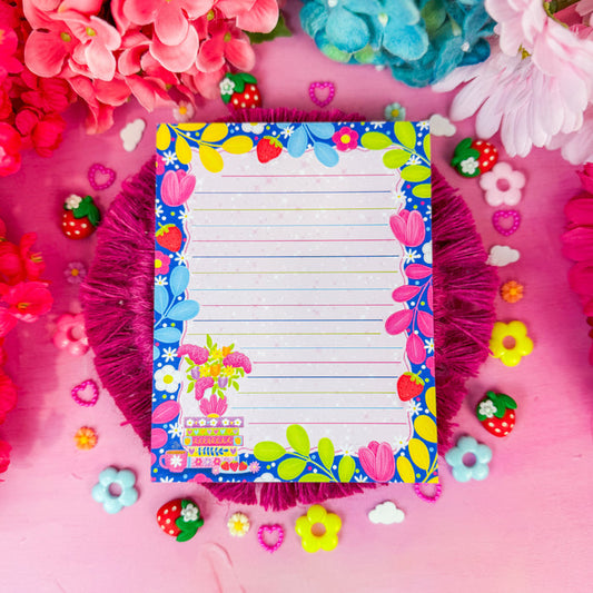 A lined notepad with a colorful floral border sits on a pink background surrounded by artificial flowers, small decorative candies, and a magenta fringed mat.